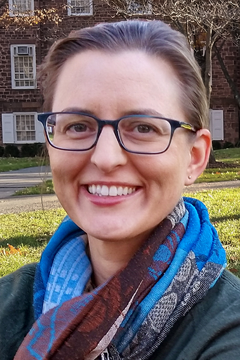 A headshot of Dr. Jaime Ahlberg wearing glasses and a blue and purple scarf. They are smiling and their hair is pulled back.