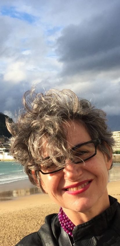 A photo of Nancy Rose Hunt at the beach and below a sky of gray clouds. She is wearing a purple beaded necklace and black glasses. She is also wearing red lipstick and smiling at the camera.