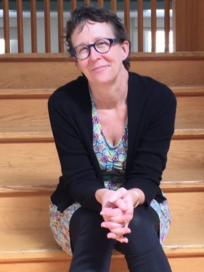 A photo of Trysh Travis seated on a staircase. She is wearing black tights, a patterned dress, and a black cardigan, with her hands clasped and resting on her knees. She is also wearing glasses, and is smiling at the camera.